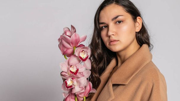 Seductive blonde woman in brown jacket posing in studio on grey background.elegant model in pastel casual spring outfit. Beautiful girl with healthy skin and branch orchid. 16:9 panoramic format. - Φωτογραφία, εικόνα
