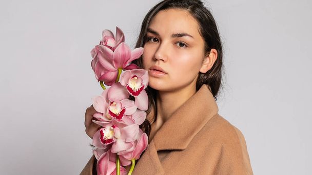 Seductive blonde woman in brown jacket posing in studio on grey background.elegant model in pastel casual spring outfit. Beautiful girl with healthy skin and branch orchid. 16:9 panoramic format. - 写真・画像