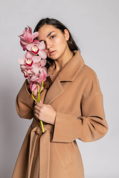 Seductive blonde woman in brown jacket posing in studio on grey background. Fashion portrait of elegant model in pastel casual spring outfit. Beautiful girl with healthy skin and branch orchid - Φωτογραφία, εικόνα