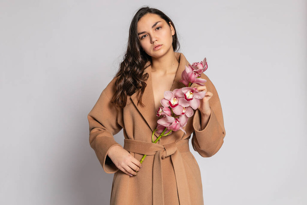 Seductive blonde woman in brown jacket posing in studio on grey background.elegant model in pastel casual spring outfit. Beautiful girl with healthy skin and branch orchid. 16:9 panoramic format. - 写真・画像