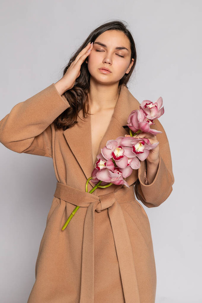 Seductive blonde woman in brown jacket posing in studio on grey background. Fashion portrait of elegant model in pastel casual spring outfit. Beautiful girl with healthy skin and branch orchid - 写真・画像