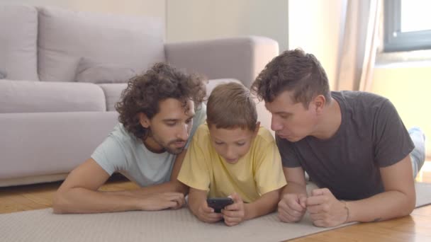 Skupiony chłopiec leżący na podłodze w pobliżu ojców i korzystający z aplikacji na komórkę - Materiał filmowy, wideo