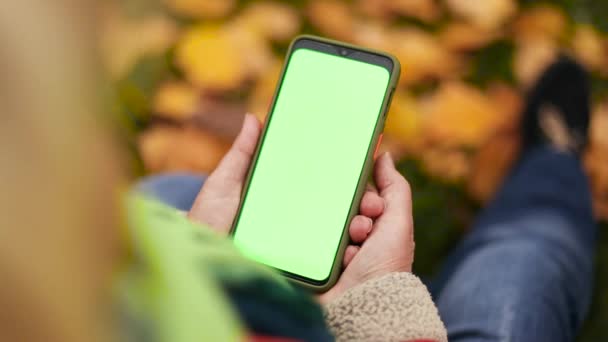 Primer plano de una mujer sosteniendo la pantalla verde del teléfono móvil al aire libre. Mujer rubia se sienta en la hierba en el parque junto a las hojas caídas - Imágenes, Vídeo