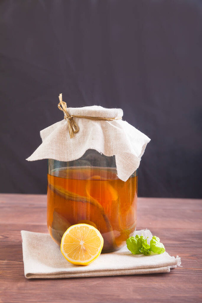 Kombucha in a glass jar, lemon and a mint leaf on a wooden table on a black background. Fermented drink. Healthy food concept. - Фото, изображение