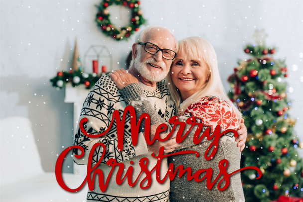 šťastný starší pár objímání při pohledu na kameru v blízkosti veselých vánočních nápisů a dekorací na rozmazaném pozadí - Fotografie, Obrázek