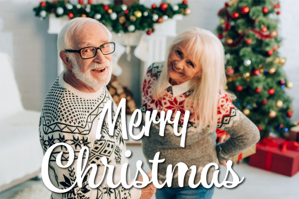 vrolijk senior man en vrouw op zoek naar camera in de buurt vrolijk kerstmis belettering en decoraties op wazig achtergrond  - Foto, afbeelding