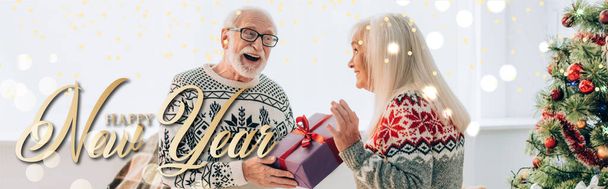 podekscytowany starszy mężczyzna prezentuje prezent dla szczęśliwej żony w pobliżu szczęśliwego Nowego Roku liternictwo, sztandar - Zdjęcie, obraz