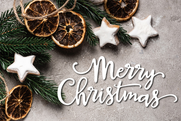 Top view of pine branch with dried orange pieces and cookies near merry christmas lettering on grey background - Foto, imagen