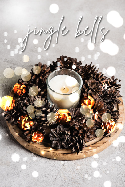 Scented candle with pine cones on wooden plate near jingle bells lettering on grey background - Fotó, kép