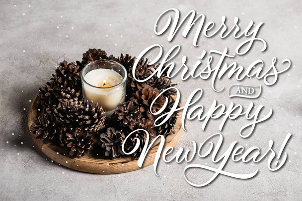 Scented candle with pine cones on wooden plate near merry christmas and happy new year lettering on grey background - Fotoğraf, Görsel