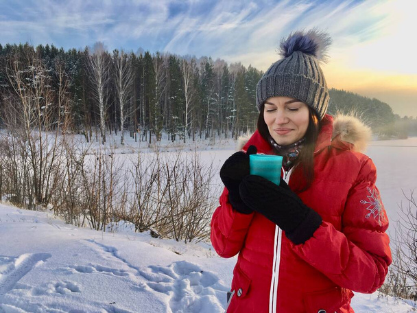 Piękna dziewczyna w zimowej kurtce z kubkiem w zimie na łonie natury. Dziewczyna w kapeluszu i czerwonej, ciepłej kurtce. Rozgrzewa ręce i pije gorącą herbatę albo drinka.  - Zdjęcie, obraz