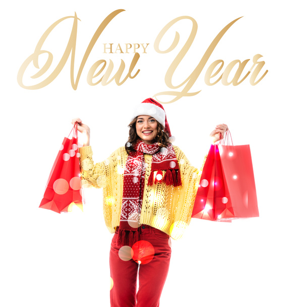 pleased woman in santa hat and scarf with ornament holding red shopping bags near happy new year lettering on white  - Фото, изображение