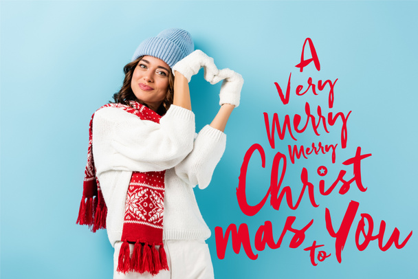 heureux jeune femme en écharpe chaude, gants et chapeau montrant signe de coeur près d'un très joyeux Noël à vous lettrage sur bleu - Photo, image