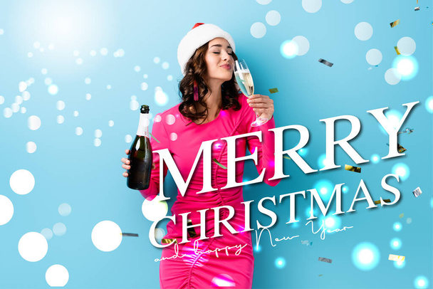 young woman in santa hat holding bottle and drinking champagne near confetti and merry christmas, happy new year lettering on blue - Photo, Image