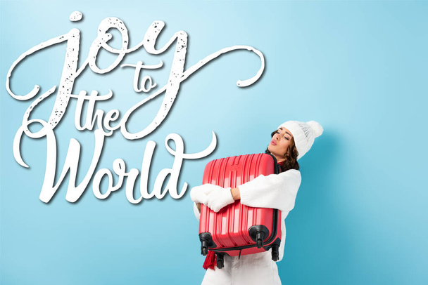 young woman in winter outfit carrying heavy suitcase near joy to the world lettering on blue - Photo, Image