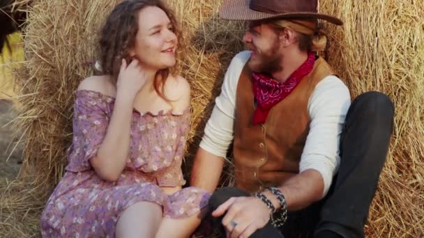 Una joven pareja se sienta en el heno de la granja. Feliz vaquero sonriente y mujer pasar tiempo juntos - Imágenes, Vídeo