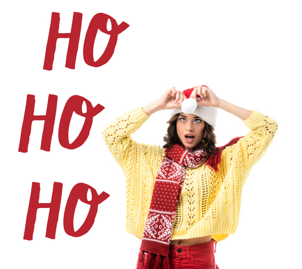 überrascht junge Frau mit Schal berührt Weihnachtsmütze in der Nähe ho ho ho Schriftzug auf weiß - Foto, Bild