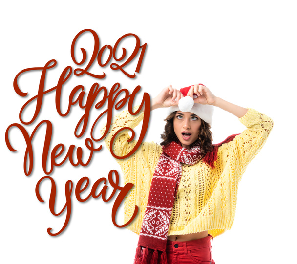surprised young woman in scarf touching santa hat near 2021 happy new year lettering on white - Photo, Image