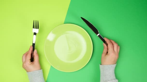 dvě ženské ruce drží kovový nůž a vidličku, uprostřed je prázdný kulatý zelený talíř, horní pohled - Záběry, video