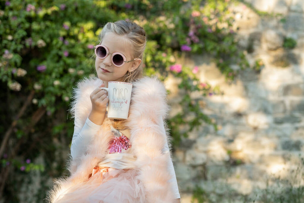 Menina de sete anos de idade, loira com cabelos longos, usando óculos de sol redondos e um colete de pele rosa, em um passeio no parque está sentado em um banco com uma xícara de chá na mão - Foto, Imagem