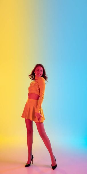 Beautiful seductive girl in fashionable, romantic yellow outfit on bright gradient yellow-blue background in neon light - 写真・画像