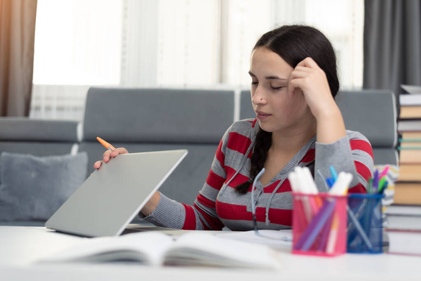社会的距離のある学生の概念、家庭での教育。高校生の女の子はノートパソコンを持って家で宿題をしている。ティーンスタディホームザ学校が閉鎖中コロナウイルスCovid-19. - 写真・画像