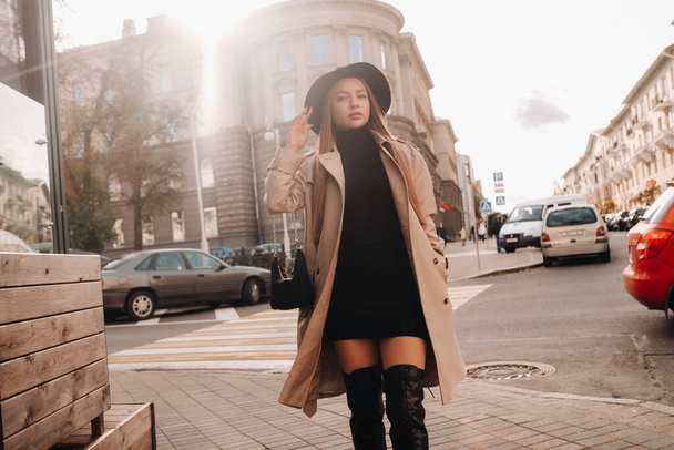 都会の通りの黒い帽子のベージュのコートを着たスタイリッシュな若い女性。女性のストリートファッション。秋服｜Urban Style. - 写真・画像