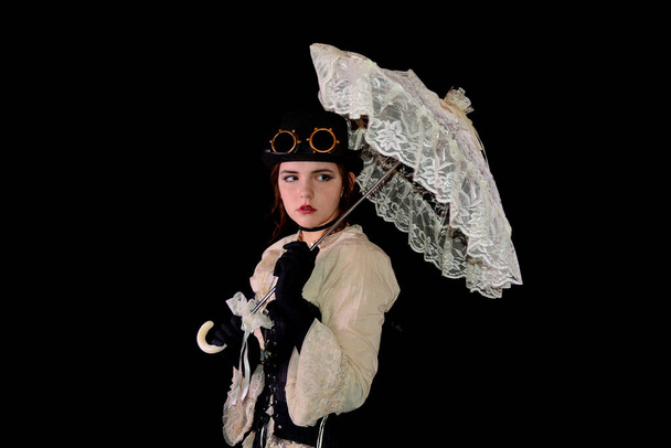 Young girl in steam punk look, holding white open umbrella made of lace, standing in front of a black background - Photo, Image