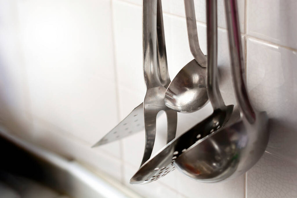 Une écumoire, deux fourchettes et deux louches en métal accrochées au mur de la cuisine carrelée. Ustensiles de cuisine en métal - Photo, image
