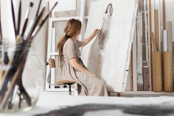 Foto de interior de una joven artista sentada frente al lienzo y pintando en un estudio blanco brillante con ropa bohemia chic con un frasco de pinceles en primer plano enfocado en el fondo - Foto, Imagen