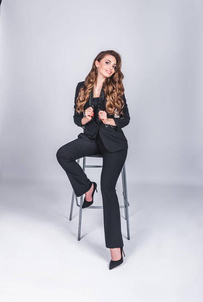 A beautiful woman in a black business suit with a make-up and long hair sits on a high chair on a white background in the studio. Working and business woman concept - 写真・画像
