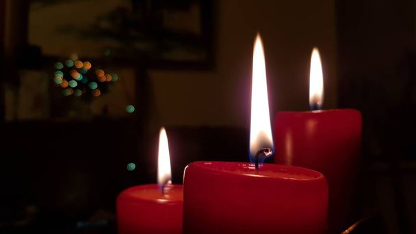 Három piros gyertya oldalnézet. Gyertyafényes lángok égő wickes közelkép. Gyertyatartók izzó tüze a sötétségben. Karácsonyi gyertyák elmosódott színes karácsonyi fények a háttérben. - Fotó, kép