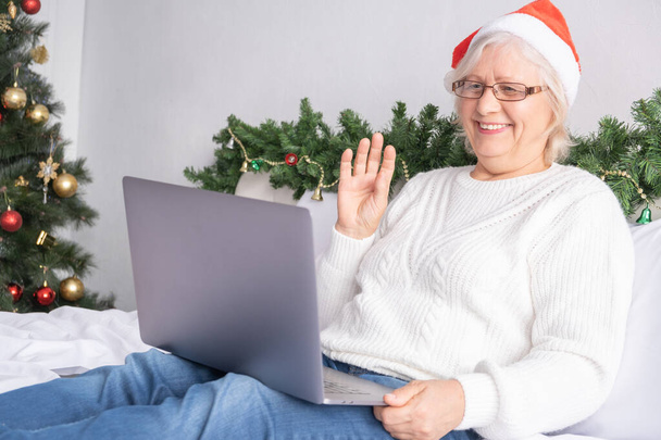 Salutations en ligne de Noël. heureuses femmes âgées dans le chapeau de Père Noël à la maison dans le lit avec ordinateur portable. - Photo, image