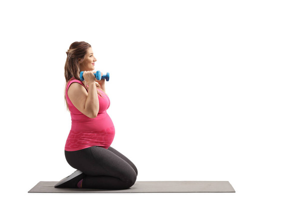 Pregnant woman kneeling and exercising with dumbbells on a mat isolated on white background - Valokuva, kuva