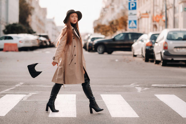 Stylish young woman in a beige coat in a black hat on a city street. Women's street fashion. Autumn clothing.Urban style. - 写真・画像