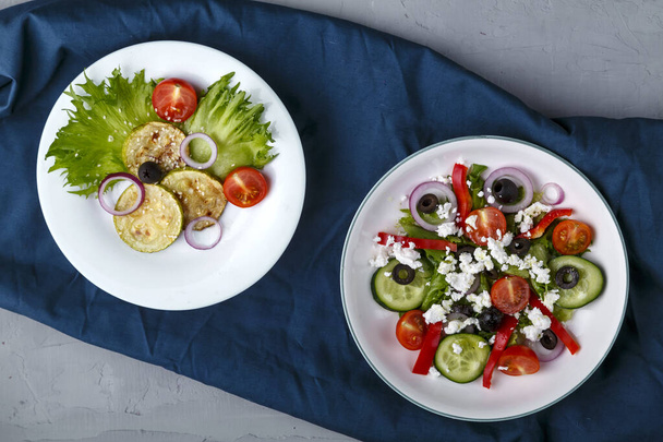Белая тарелка с жареными цуккини с помидорами черри на салате и греческий салат на голубой салфетке. - Фото, изображение