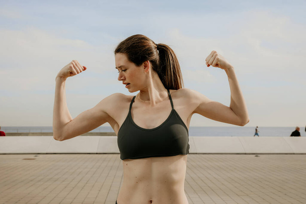 Portrait of young sporty woman showing biceps posing outdoor. - Фото, изображение
