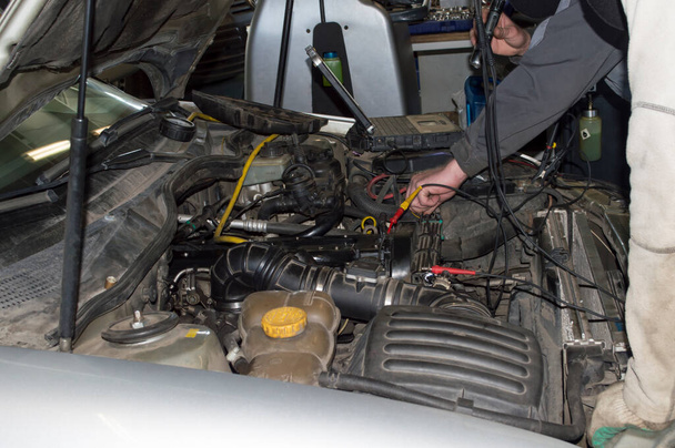 Un electricista en uniforme gris comprueba el equipo eléctrico debajo del capó del coche usando una computadora en el taller de reparación del coche - Foto, Imagen