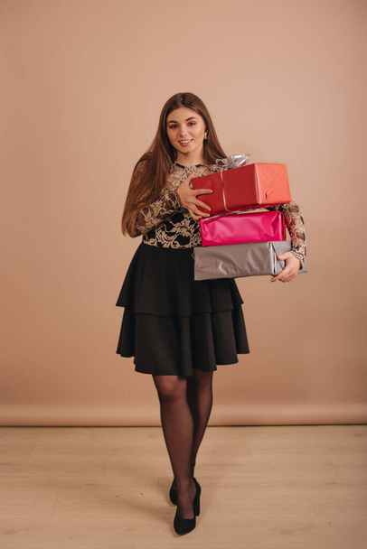 A girl with long hair, in an elegant black dress, holding many colorful gift boxes in her hands against a beige background. - Foto, Bild