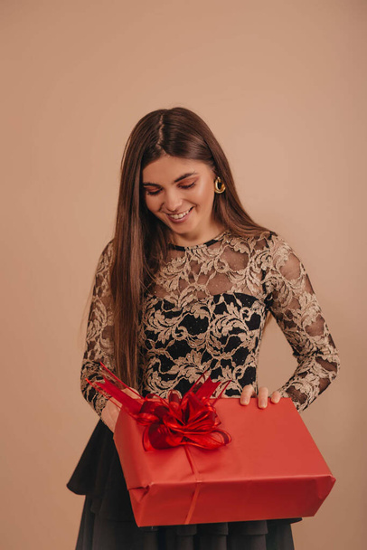 усміхнена дівчина в елегантній чорній сукні відкриває свій новорічний подарунок ізольовано на бежевому кольоровому фоні
.  - Фото, зображення