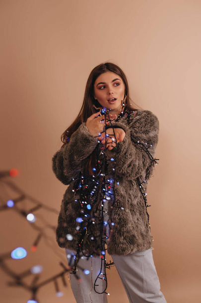 A beautiful girl in a gray fur coat is holding a luminous garland in her hands against beige color background. Glare from the garland. - Photo, image