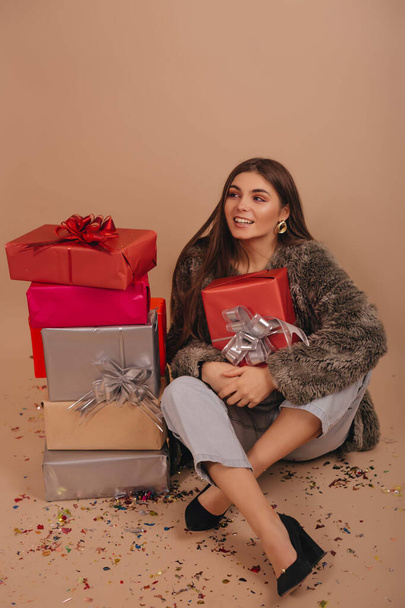 Gülümseyen kız, bej renkli arka planda izole edilmiş hediye kutularının yanında oturuyor..  - Fotoğraf, Görsel