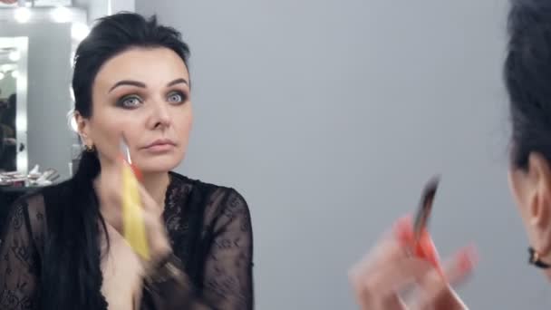 Une belle jeune femme spectaculaire dans un peignoir noir noir et décolleté applique du maquillage sur son visage avec un pinceau spécial devant un miroir - Séquence, vidéo