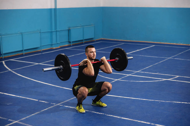 Fitter muskulöser, kräftiger Bodybuilder in Sportbekleidung, der Gewichte hebt, während er in der Sporthalle hockt. - Foto, Bild