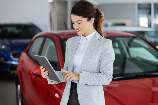 Smiling car seller in suit standing in car salon and using tablet for choosing right car. In background are many modern cars. - Photo, image