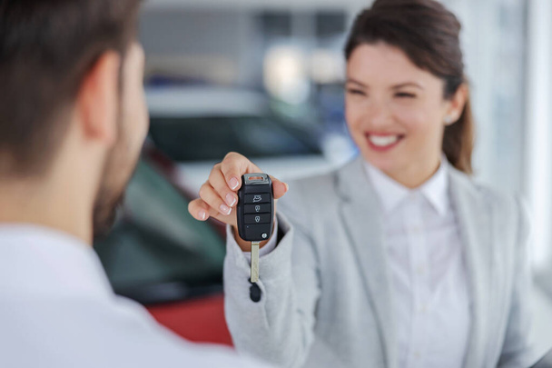 Closeup of smiling female car seller handing a keys to a male buyer while standing in car salon. Selective focus on keys. - Photo, Image