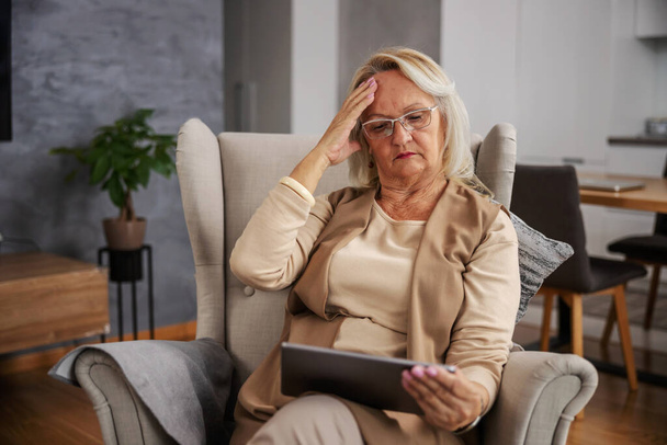Mulher idosa loira sentada em casa, com dor de cabeça e olhando para tablet para obter conselhos on-line. - Foto, Imagem
