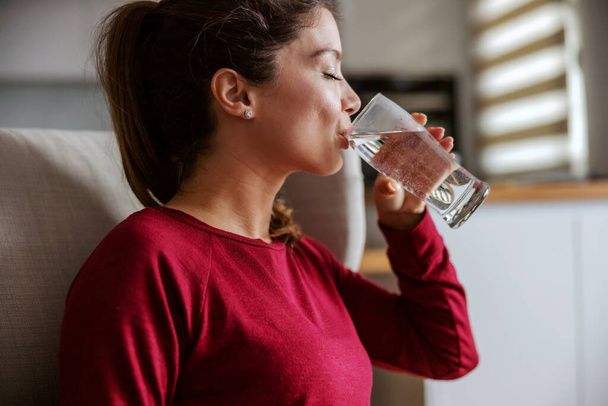 Profil młodej brunetki siedzącej w domu i pijącej wodę. - Zdjęcie, obraz
