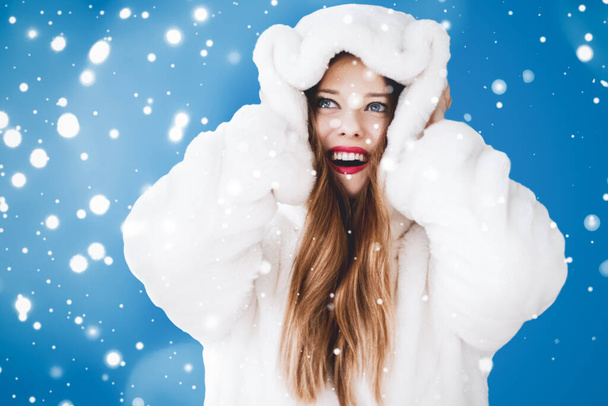 幸せなクリスマスと白いフード付きの毛皮のコートで若い女性の冬の休日の肖像画、青い背景に雪、ファッションとライフスタイル - 写真・画像