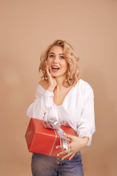 glimlach verrast blond vrouw in wit blouse holding rood gift box geïsoleerd op beige achtergrond. - Foto, afbeelding